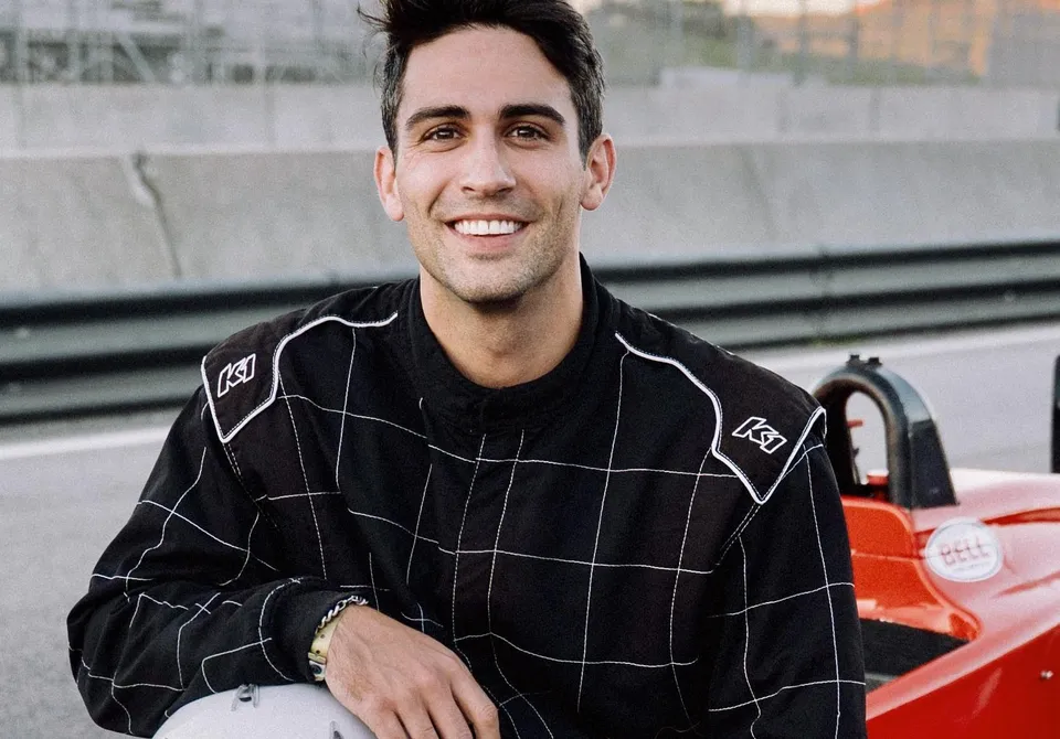 Thomas in a black race suit, smiling, next to a red open-wheel race car at a track